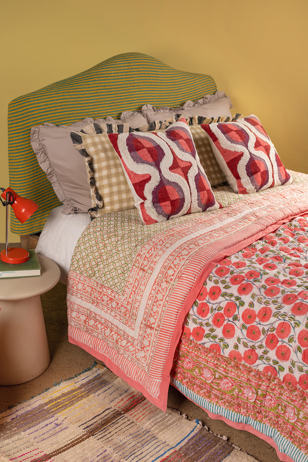 A cozy bed features a colorful striped headboard, patterned pillows, and the Aiko Cotton Quilt by Ian Snow Ltd with geometric floral design. An orange lamp and books sit on the side table against a yellow wall with a woven rug below.